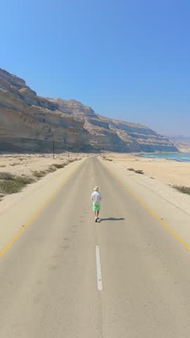 Aerial view of a young man walking down an empty coastal highway flanked by high cliffs and the sea in Salalah, Oman.