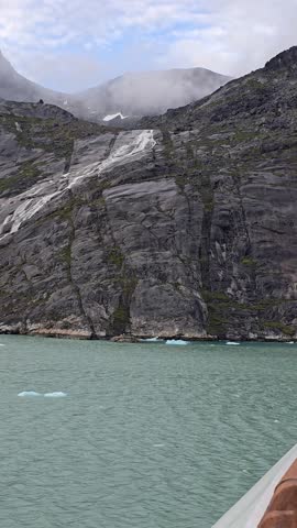 Blue Skies, Glaciers, mountains and Icebergs of Prins Christian Sund, South Greenland, August 2025. Rocky coastal terrain of Prins Christian Sund, South Greenland, partially shrouded in mist. The foreground features rugged rocks with patches of greenery, leading down to calm, dark teal water. Above, the sky is blue, contrasting with dense clouds or fog obscuring the view of bigger mountainous features behind. The scene exemplifies the raw, natural beauty typical of Greenland