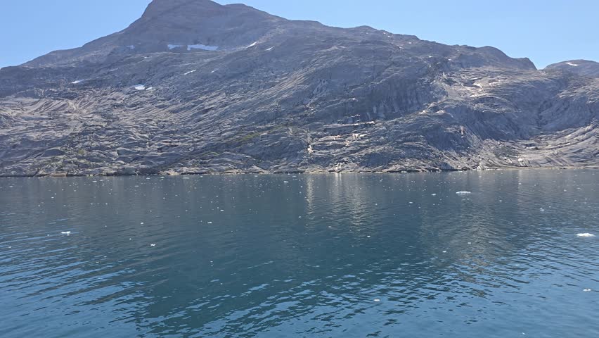 Blue Skies, Glaciers, mountains and Icebergs of Prins Christian Sund, South Greenland, August 2025. Rocky coastal terrain of Prins Christian Sund, South Greenland, partially shrouded in mist. The foreground features rugged rocks with patches of greenery, leading down to calm, dark teal water. Above, the sky is blue, contrasting with dense clouds or fog obscuring the view of bigger mountainous features behind. The scene exemplifies the raw, natural beauty typical of Greenland