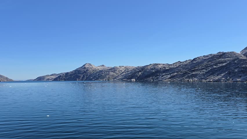 Blue Skies, Glaciers, mountains and Icebergs of Prins Christian Sund, South Greenland, August 2025. Rocky coastal terrain of Prins Christian Sund, South Greenland, partially shrouded in mist. The foreground features rugged rocks with patches of greenery, leading down to calm, dark teal water. Above, the sky is blue, contrasting with dense clouds or fog obscuring the view of bigger mountainous features behind. The scene exemplifies the raw, natural beauty typical of Greenland