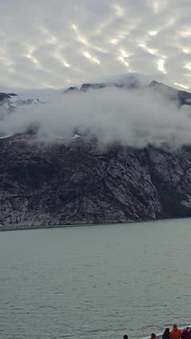 Blue Skies, Glaciers, mountains and Icebergs of Prins Christian Sund, South Greenland, August 2025. Rocky coastal terrain of Prins Christian Sund, South Greenland, partially shrouded in mist. The foreground features rugged rocks with patches of greenery, leading down to calm, dark teal water. Above, the sky is blue, contrasting with dense clouds or fog obscuring the view of bigger mountainous features behind. The scene exemplifies the raw, natural beauty typical of Greenland
