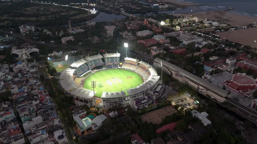 Aerial video of brightly illuminated a cricket stadium during an evening match in Chennai, India. The iconic circular stadium is surrounded by dense urban neighborhoods, with the coastline and Beach