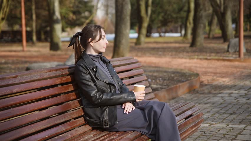 Young adult woman wearing black leather jacket and dark clothing sits relaxed on wooden bench in autumn park drinking hot coffee from disposable cup with calm expression