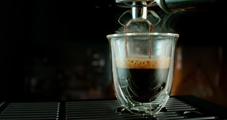 Super slow motion close-up of espresso pouring from a coffee machine into a glass cup. Rich crema forms on the surface as dark coffee flows, highlighting texture, aroma and detail of fresh coffee preparation.