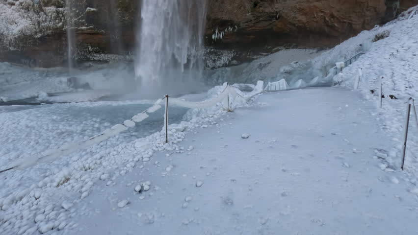 Tilting shot of large waterfall surrounded by ice in late winter in Iceland