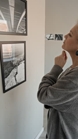 Vertical slowmo shot of mature Caucasian woman observing framed artwork on exhibition wall and abstract black sculpture on pedestal while visiting modern art gallery