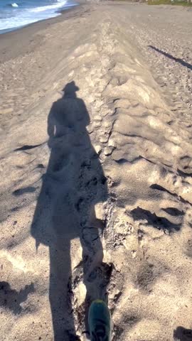 POV footage of a person walking along a sandy beach showing shadow and footsteps. Minimal coastal scene with natural textures, suitable for travel, lifestyle, and relaxation themes.