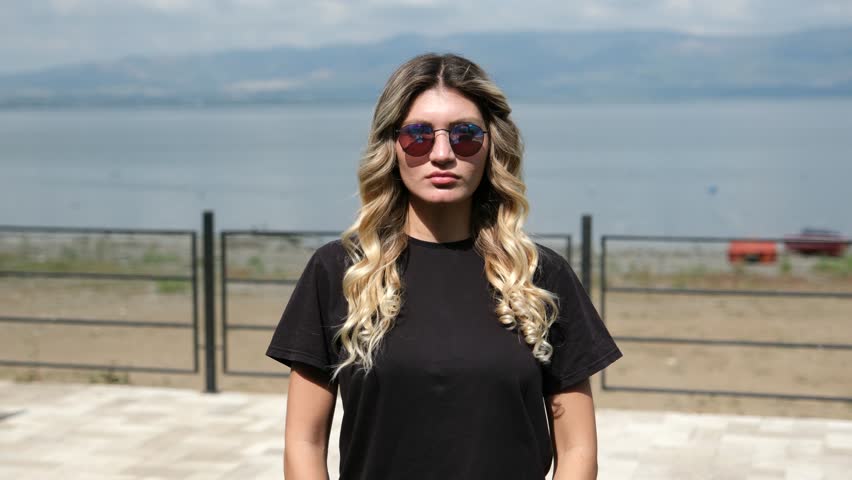 Beautiful young woman wearing sunglasses standing on a coastline by the sea and looking at camera during a bright sunny summer vacation trip at a Mediterranean beach resort