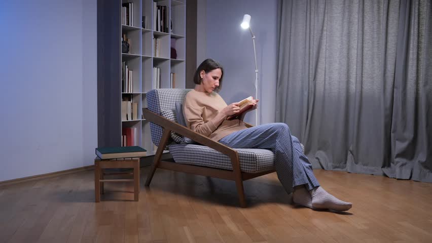 Woman relaxing in a cozy armchair at night, reading a book under a warm floor lamp in her home study, enjoying quiet, focused learning and peaceful evening downtime