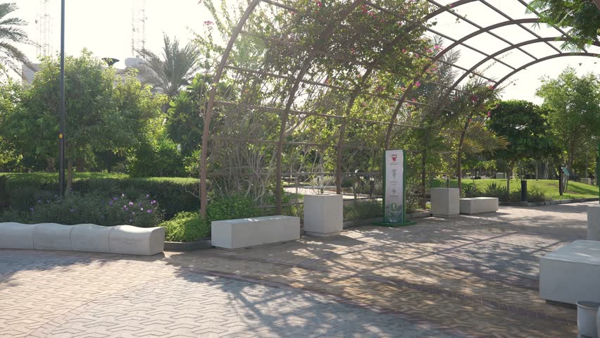 A serene park area featuring a metal pergola covered with greenery, surrounded by benches and blooming flowers under bright sunlight.