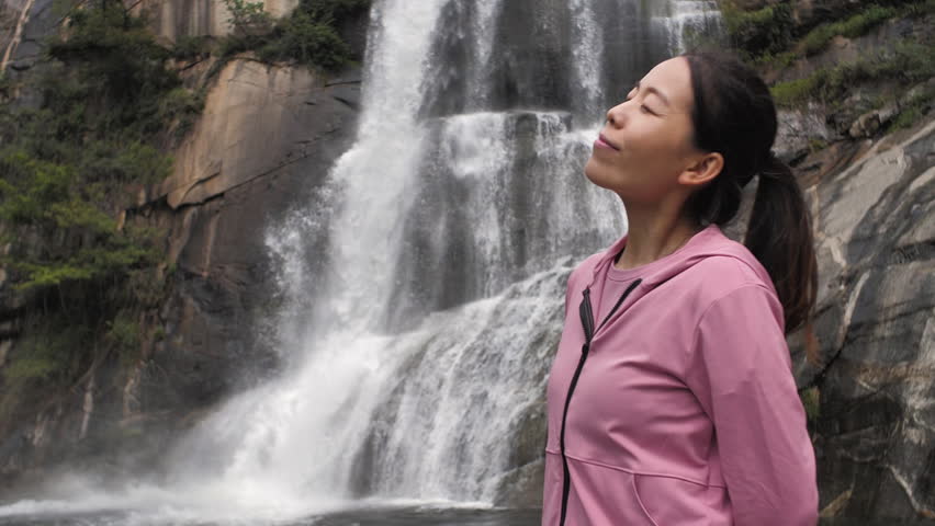 Asian Chinese women embrace nature with open arms beneath a waterfall.