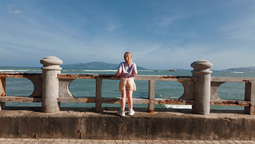 Young girl looks at ocean from stone railing by beach during daytime