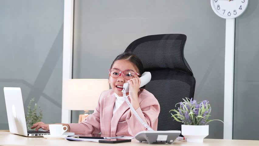A young girl wearing a pink suit talks on a telephone at an office desk