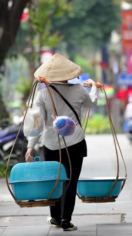 Street vendor person in conical hat walking down street in Hanoi, Vietnam carrying shoulder cooking pans , traditional local Vietnamese trader balancing stuff on shoulder pole while walk in city area.