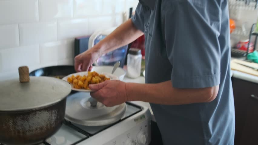 Asian Elderly Man Frying Potatoes On Stove Pan And Transferring To Plate Family Cook Searing Golden Pieces With Spatula Under Bright Kitchen Light, Sizzling Aroma And Careful Motion Near Tiled