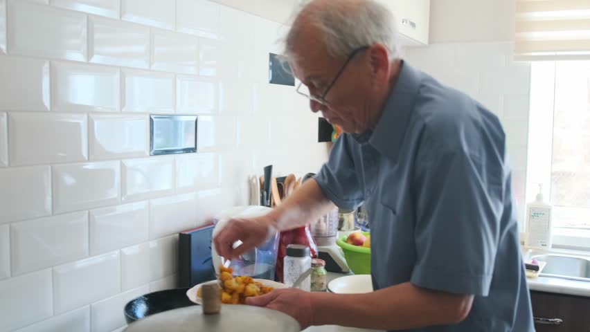Asian Elderly Man Frying Potatoes On Stove Pan And Transferring To Plate Family Cook Searing Golden Pieces With Spatula Under Bright Kitchen Light, Sizzling Aroma And Careful Motion Near Tiled