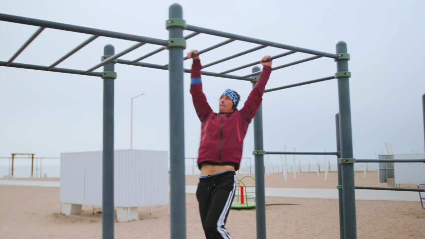 Male Athlete Performs Swing At Seaside Park, Caucasian Male Excercises On Playground Equipment Near Shoreline, Man Actively Uses Monkey Bars During Outdoor Workout At Beachside Recreational Area