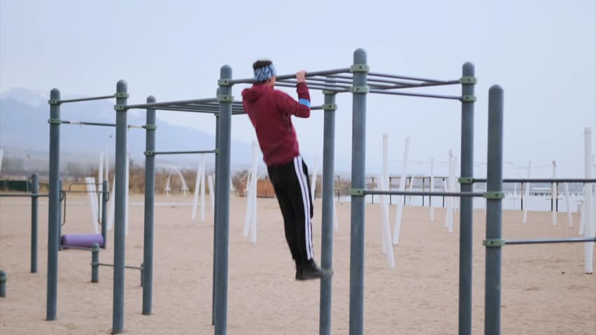 Caucasian Man Approaching Outdoor Bars On Beach, Purposeful Stride Toward Calisthenics Frame, Layered Jacket And Headband, Empty Seafront Training Area, Focused Preparation For Hanging Routine