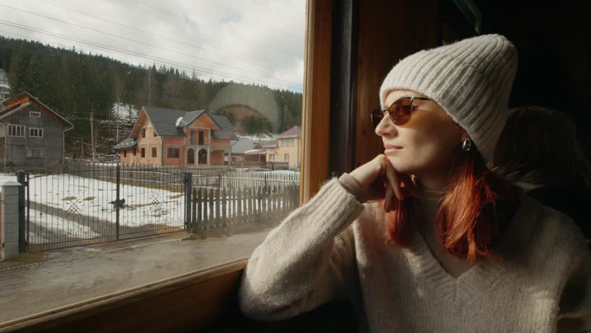 A woman rides on an old train, looking out the window. She enjoys the changing views of nature and landscapes while traveling through Romania.