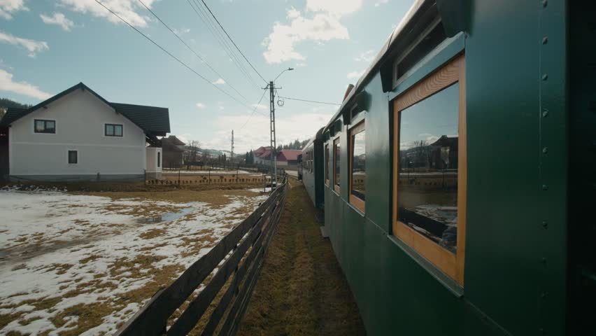 The Mocanita steam train moves through the villages in Bucovina, Romania. It passes by houses and fields under a clear sky during the day.