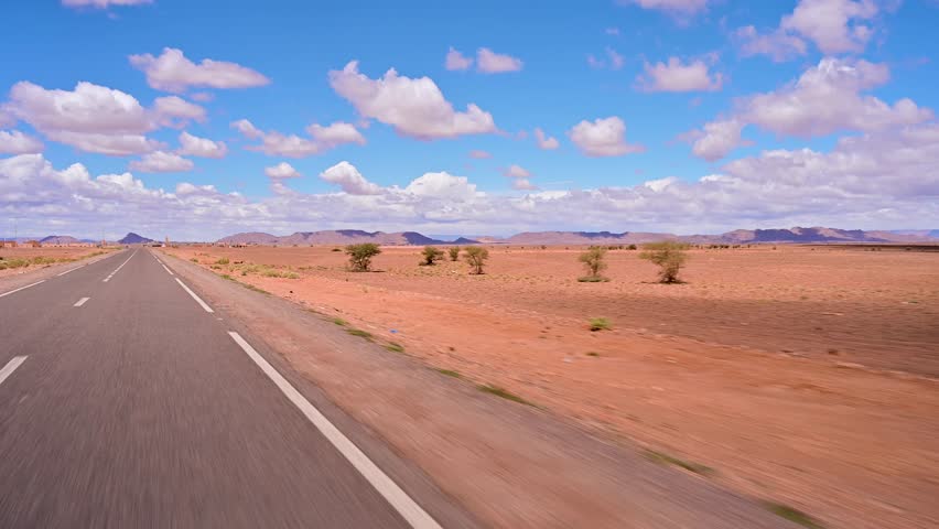 Point of view shot driving on a long, empty asphalt road through the arid landscape of the Sahara Desert under a blue sky in Morocco