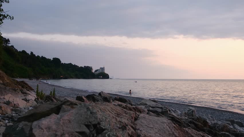 Scenic view of a grey pebble coastline with large rocks in the foreground, calm sea water, and a modern white hotel building situated on a lush green hill under an overcast sky.