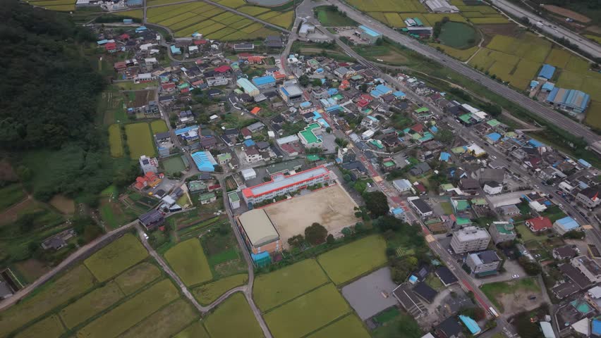 This high-angle aerial perspective captures a colorful South Korean village characterized by traditional and modern houses with brightly painted roofs. In the foreground, a local primary school with a distinct red roof and dirt playground stands next to lush rice paddies, while a highway borders the village in the background. Concept of rural development and community life.