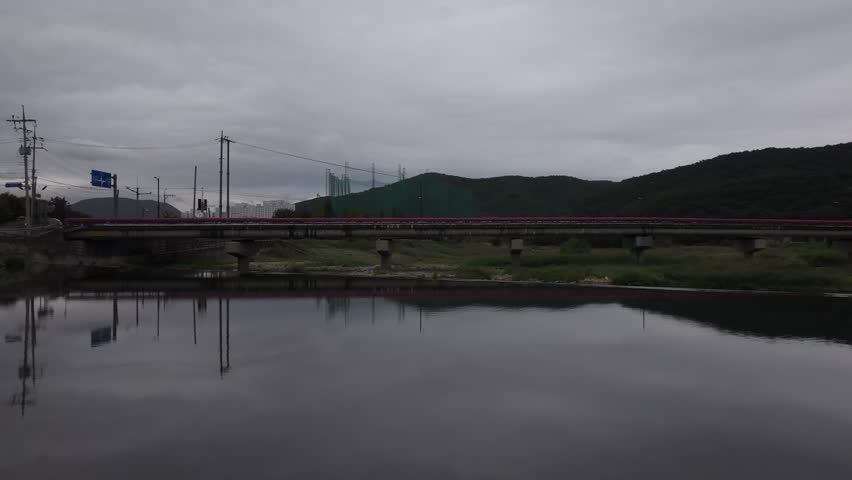 A low-angle shot shows a sturdy concrete bridge spanning across a shallow, rocky riverbed with sparse water puddles. The bridge railing is adorned with a dense, continuous row of vibrant pink flowers, creating a sharp contrast against the overcast grey sky and the muted tones of the landscape. In the background, lush green hills are visible under a cloudy sky, while the foreground is dominated by smooth river stones. Concept of urban landscaping and environmental beauty.