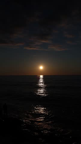 Vertical screen sun setting over sea with bright reflection on water silhouette of person near shore dramatic evening sky calm waves and peaceful atmospheric mood