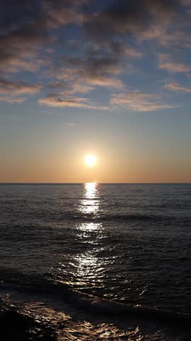 Vertical screen sun setting over sea horizon reflection on water gentle waves and soft clouds peaceful coastal scene with warm evening light and tranquil mood