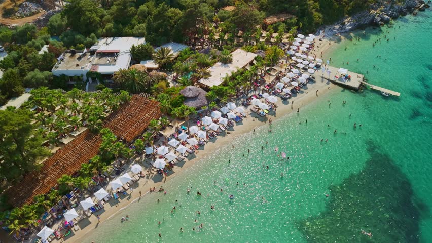 Aerial view of a luxury beach club with white umbrellas, palm trees, and a wooden pier on a clear turquoise sea. La Scala Beach, Thasos, Greece
