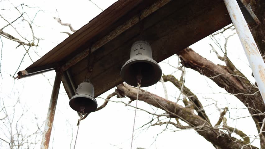 Old metal church bells hanging under rusty roof with ropes traditional religious objects outdoor setting with bare tree branches and vintage atmosphere