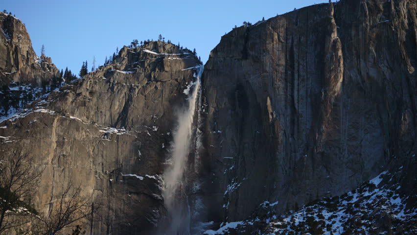 Winter waterfall plunging from granite cliffs Yosemite fall sends misty plume against sheer rock faces icy ledges and shadowed canyon creating dramatic winter panorama, Yosemite National Park