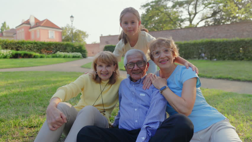 Senior people sitting on grass with family, elderly man and two older women embraced by young girl in sunny neighborhood park, casual clothing, warm smiles and affectionate hugs, relaxed