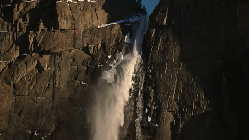 Warm sunset light on waterfall descending granite wall, amber highlights and long shadows, dramatic textures and evening calm, cinematic late light emphasizing vertical flow. Yosemite National Park