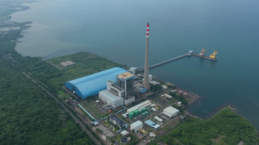 Drone footage capturing an expansive view of a coal power plant on the coast. The industrial facility features a tall chimney and a pier