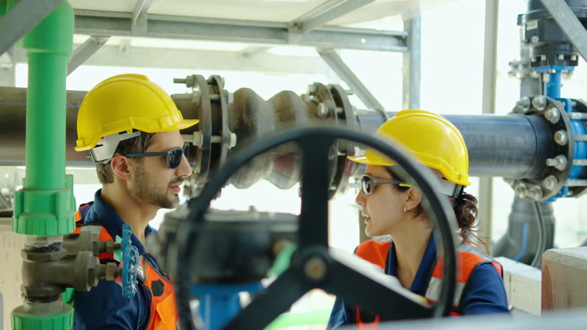 Professional male and female engineers in safety gear inspect industrial pipes and valves at a plant while using a walkie-talkie for effective communication and maintenance coordination tasks.