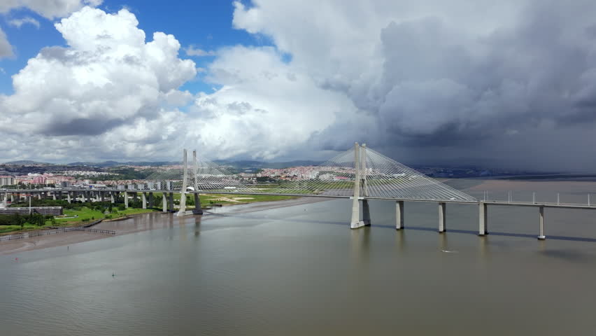 Aerial view of the Vasco da Gama Bridge in Lisbon, Portugal, spanning the Tagus River. A dramatic sky shows a contrast between blue sky with white clouds and dark, approaching storm clouds