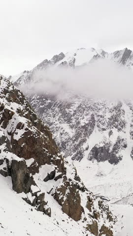 Vertical shot of snowy mountain landscape with rocky cliffs and low clouds creating dramatic foggy atmosphere. Alpine terrain covered with snow in cold weather with minimal composition and copy space