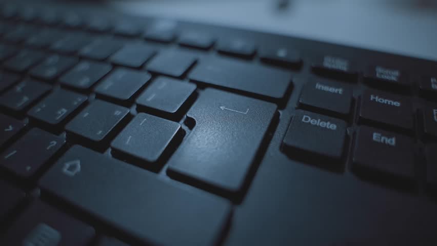 Extreme close up on finger pressing on enter or return key on computer keyboard. Person clicking on button to confirm or execute command or submit data. Dark room or office desk. Hacker or programmer