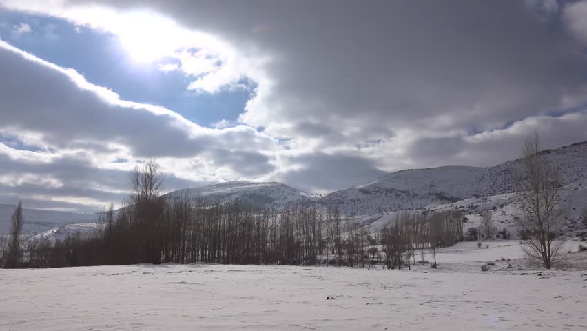 Timelapse snowy Iranian Plateau hills and leafless trees under winter clouds across frozen land. Fast moving sky sweeps above barren Persian uplands, serene ridges, icy ground, remote scenery.