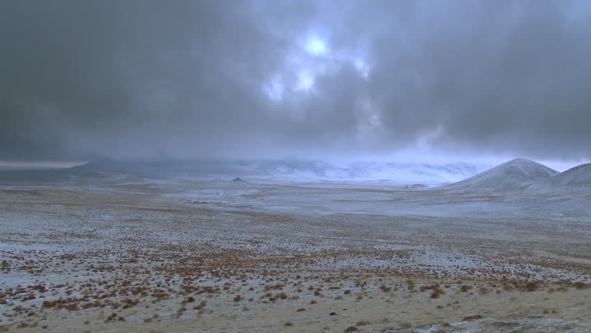Mongolian steppe in winter with snow covered plains, mountains, and fast moving clouds in timelapse. Fixed camera records frozen grassland beneath stormy sky as icy weather sweeps across remote Asia.