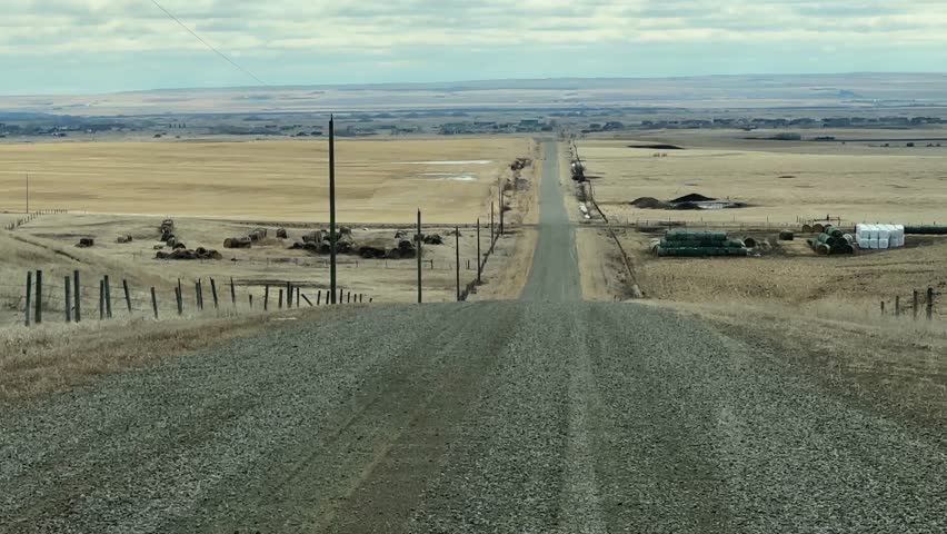 Driving POV along dusty backcountry road surrounded by open prairie fields and rural landscape