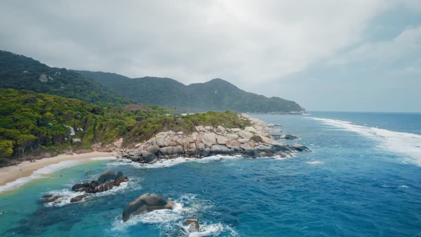 Aerial drone shot captures the breathtaking beauty of Tayrona National Park, Colombia. Turquoise waters, sandy beaches, lush jungle mountains, and a traditional hut invite tourists to explore this vibrant Caribbean paradise.