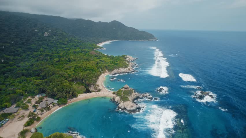Aerial drone footage showcases the breathtaking beauty of Tayrona National Park in Santa Marta, Colombia. Pristine turquoise waters, golden sandy beaches, lush tropical forests, and waves crashing against rocky shores create a picturesque coastal landscape. People enjoy the idyllic beach.