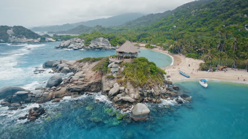 Stunning aerial view of Cabo San Juan del Guia beach in Tayrona National Park, Colombia. Features a rustic hut on a rocky outcrop, turquoise Caribbean Sea, white sand, and lush tropical jungle. A pristine travel destination.