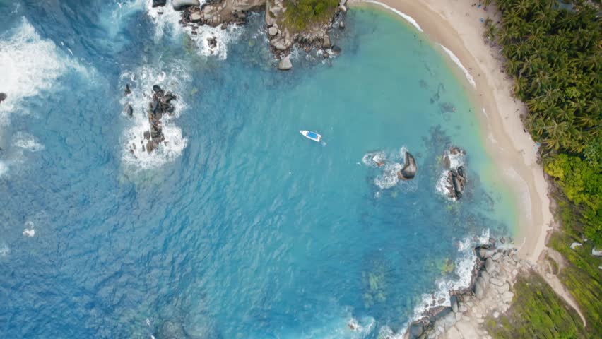 Aerial drone shot showcasing the stunning tropical paradise of Tayrona Natural Park in Santa Marta, Colombia. Turquoise waters meet sandy beaches and lush palm trees, with waves crashing on rocky shores and a small boat anchored in the bay.