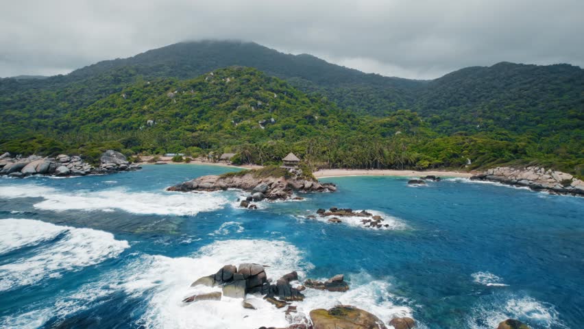 Aerial drone shot captures the breathtaking beauty of Tayrona National Park in Santa Marta, Colombia. Turquoise Caribbean sea meets lush green mountains, palm-fringed beaches, and rocky islets with a traditional hut.