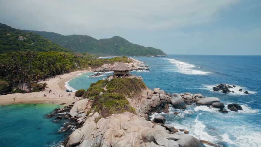 Panoramic aerial view captures the breathtaking El Cabo San Juan beach in Tayrona National Park, Santa Marta, Colombia. Turquoise Caribbean Sea meets golden sands, framed by lush jungle mountains and a distinctive thatched-roof hut on a rocky peninsula. People enjoy the vibrant tropical paradise, swimming and relaxing on the beautiful shore. Waves crash against the rugged coastline, highlighting the natural beauty of this famous destination.