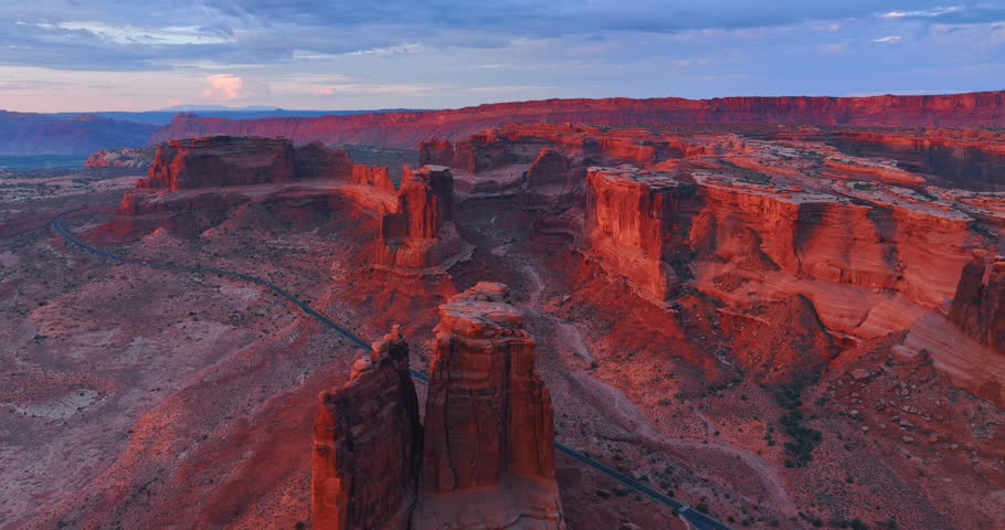 Setting sun lighting the red rocks of amazing Arches National Park, Utah, USA. Few cars ride by the highway crossing the scenery. Aerial view.
